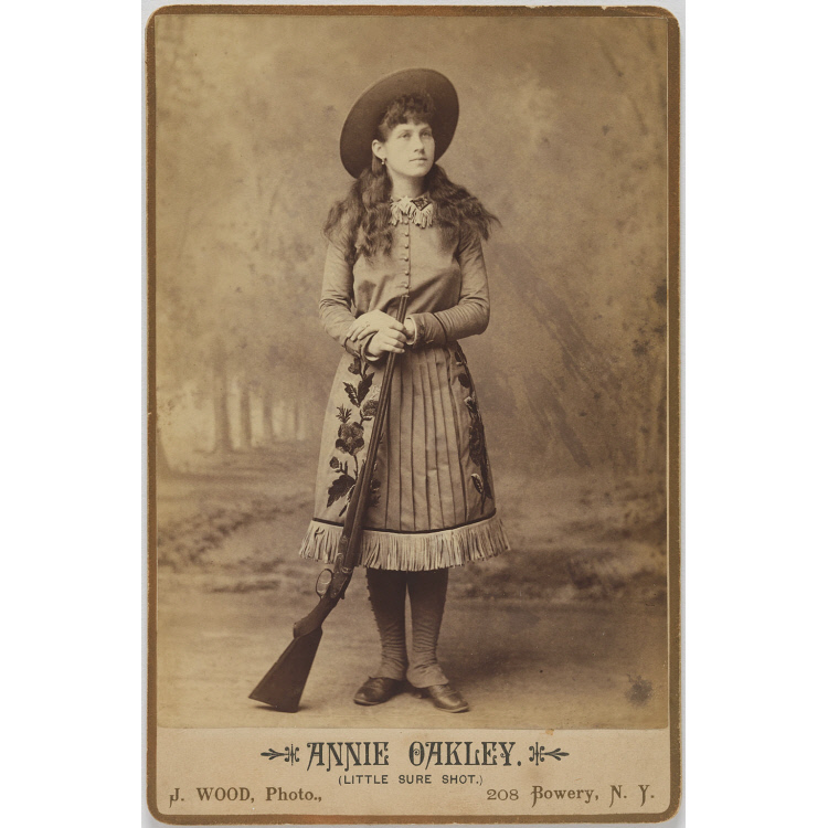 Photograph of a woman in western garb with a gun leaning against her leg