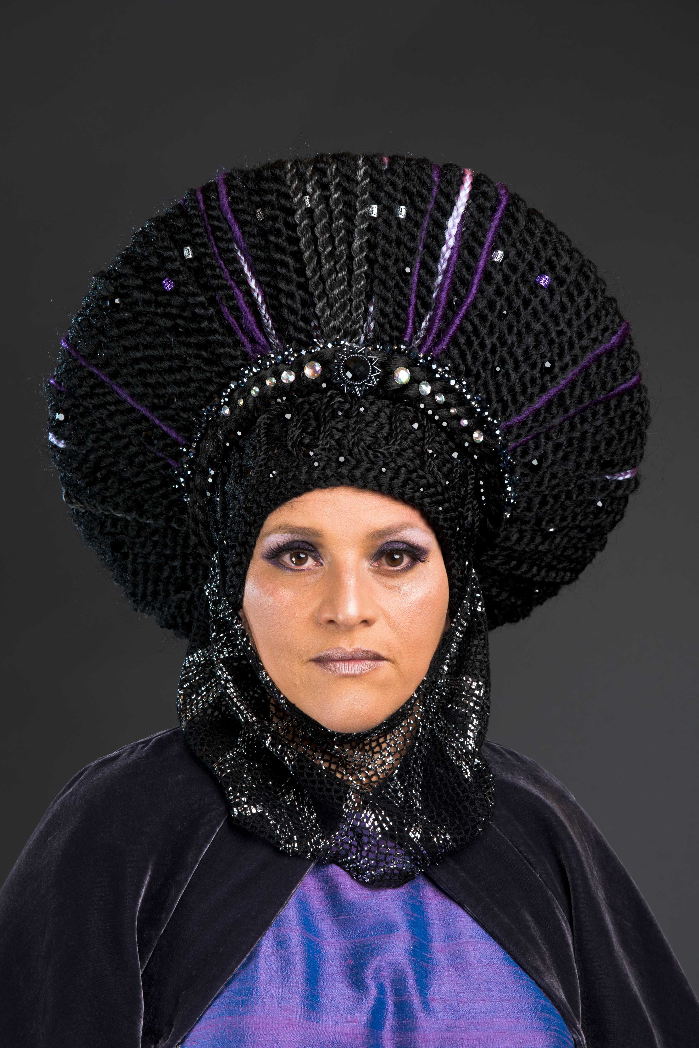 A head shot of a woman wearing a purple robe and headdress looking straight into the camera