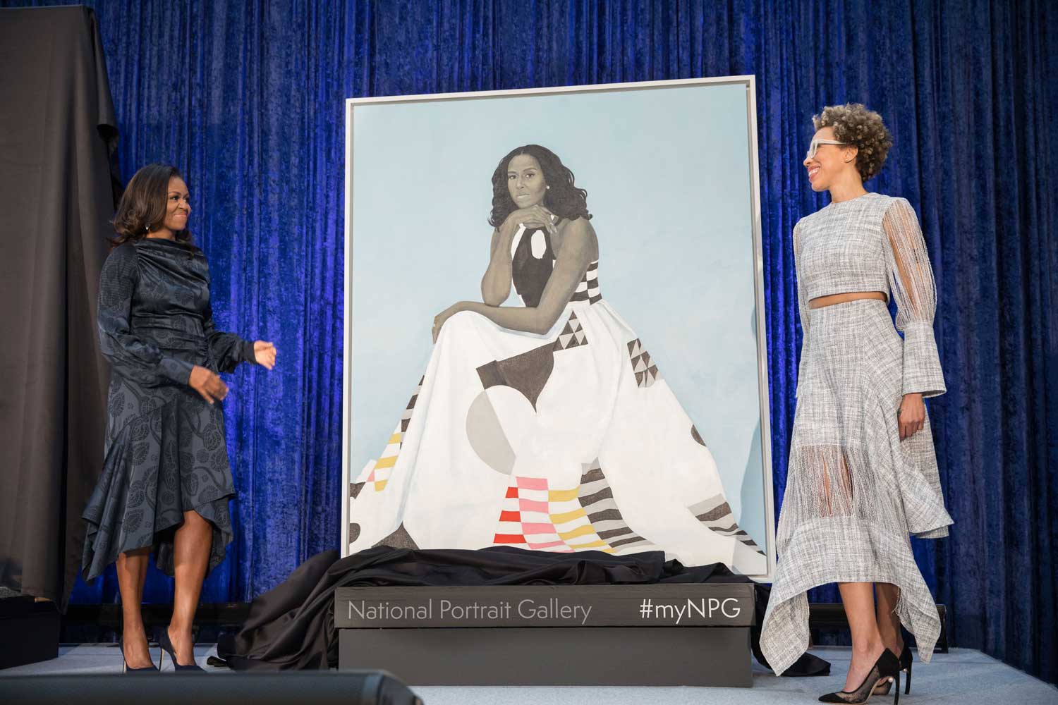 Two women standing on a stage next to a large portrait of a woman