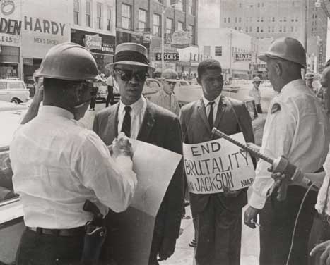 Black and white photo of Roy Wilkins and Medgar Evers, dressed in suits and being confronted by two police officers