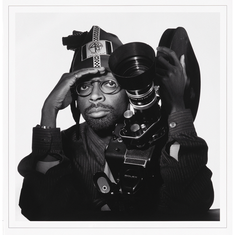 Black and white photo of Spike Lee holding film camera, with his right hand shielding his eyes as if from a bright sun