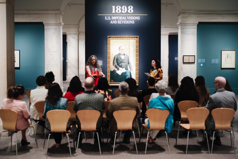 Two women discuss a portrait before a seated audience.