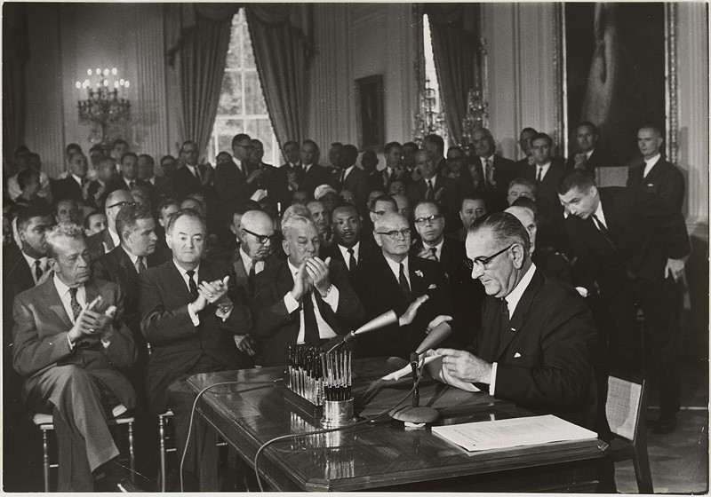 Una imagen en blanco y negro de un hombre sentado en un escritorio firmando un trozo de papel con muchos hombres aplaudiendo detrás de él