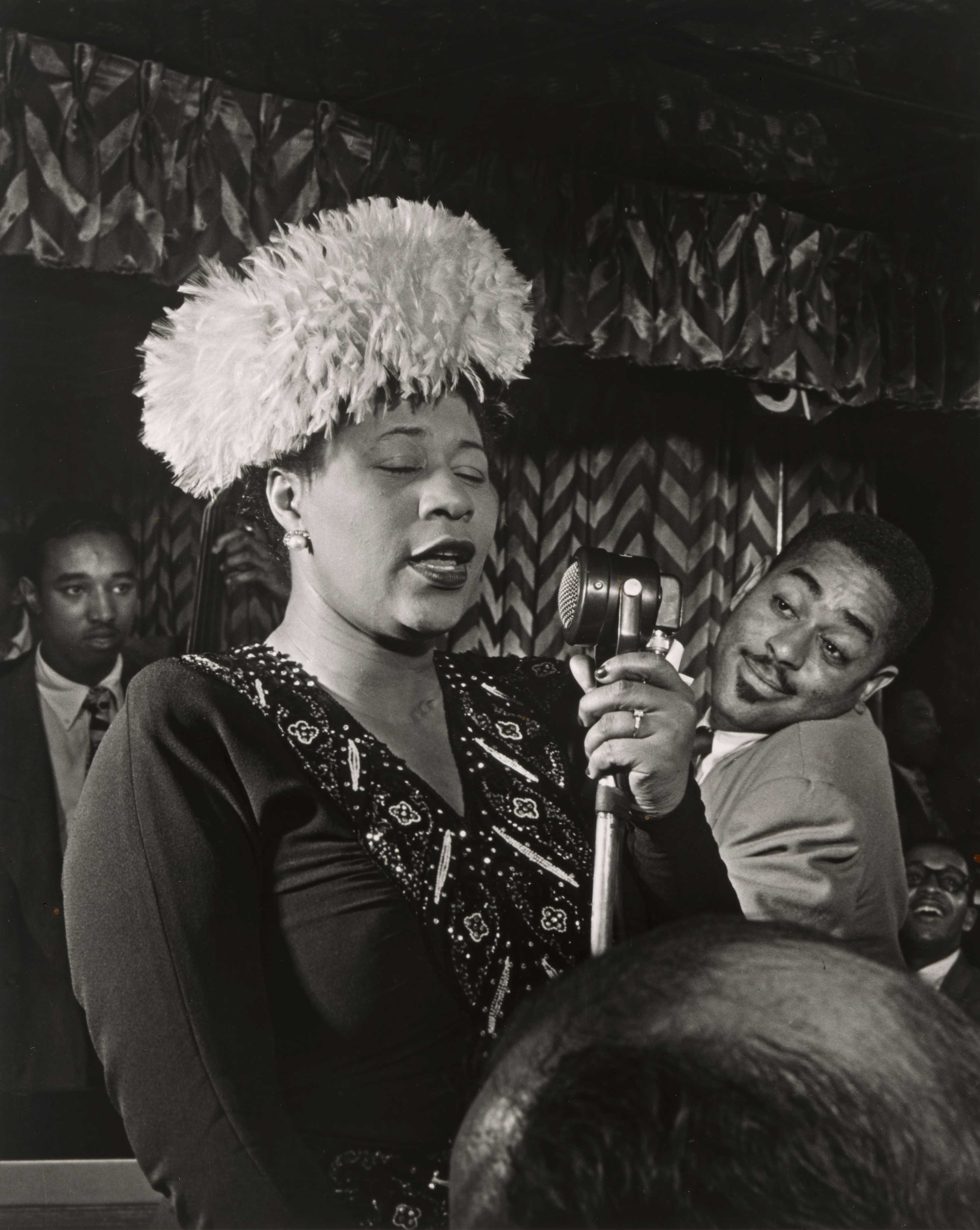 Black and white image of a woman singing into a microphone and wearing a hat