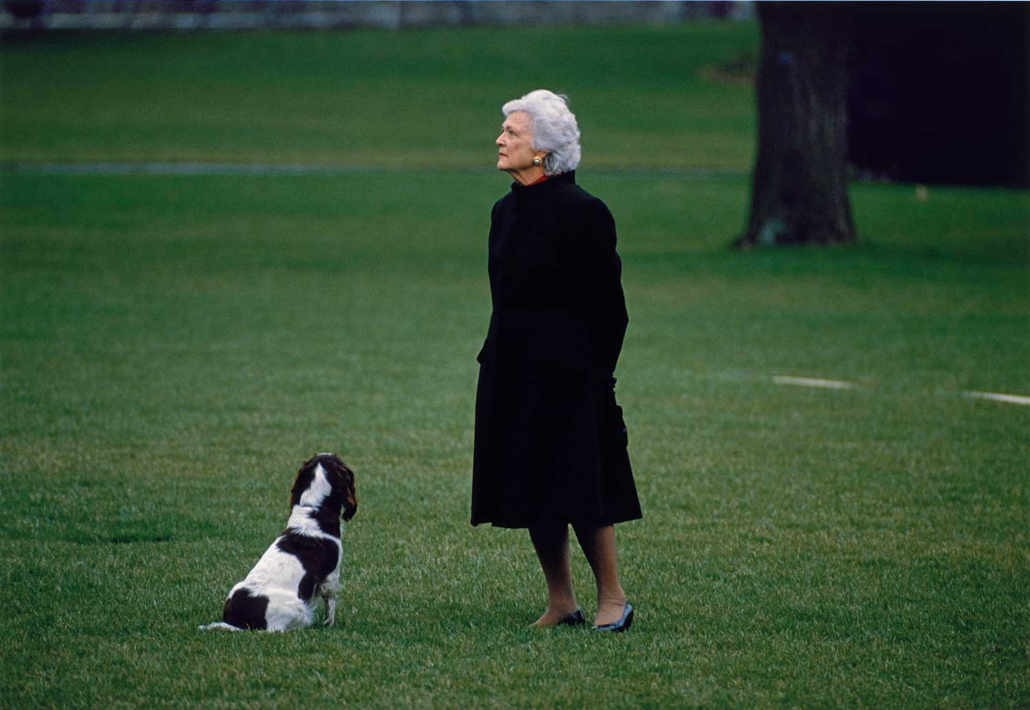 A photograph of a woman wearing a black dress and standing next to a dog outside