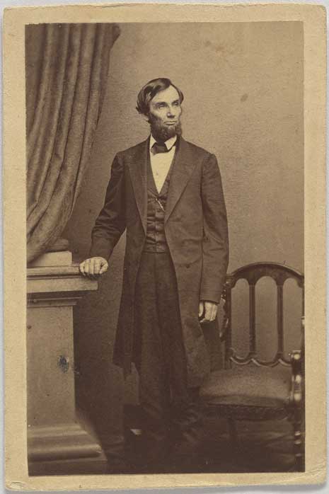 Blog_lincoln_gettysburg Black and white photo of Abraham Lincoln, standing next to a chair, with his right hand posed on a pedestal.