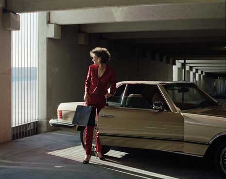 Color photo of a woman in a red business suit standing next to a car with the door open, in a parking garage.