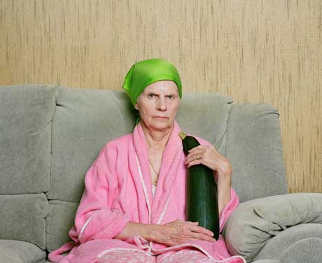 Color photo of an elderly woman in a pink bathrobe, holding a zucchini, sitting on a gray couch.