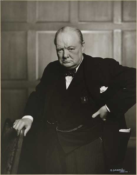 Black and white photo of Sir Winston Churchill, waist up, in a black suit and bow tie, his right hand on a chair.