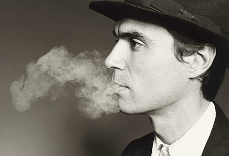 Black and white photo of David Byrne, smoking, wearing a dark suit and hat.