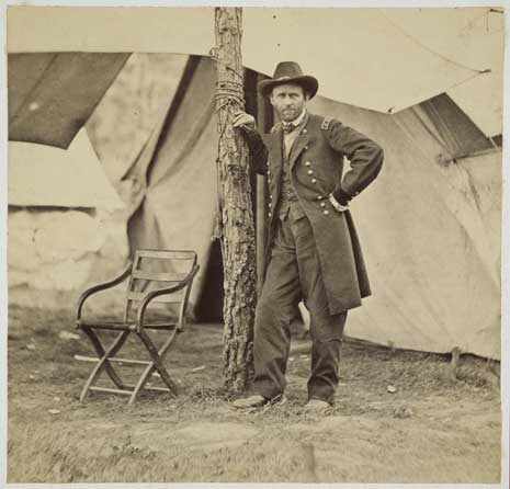 Blog_grant_by_brady Black and white print of Ulysses S. Grant standing in military uniform, leaning on a tree, in a military camp.