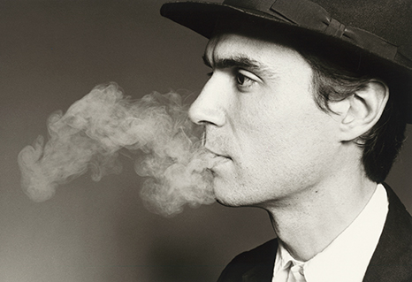 Blog_david_byrne Black and white headshot photo of David Byrne smoking, in a suit and tie with a hat.