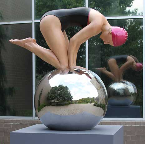 Sculpture of a swimmer kneeling on a metal ball, mounted on a pedestal.