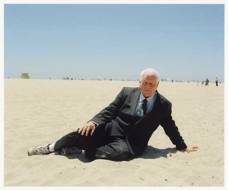 Color photo of an elderly man in a black business suit on a beach sitting in the sand.