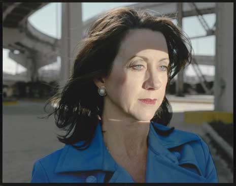 Color photo of a woman from the shoulders up wearing a blue jacket, standing beneath a highway overpass.
