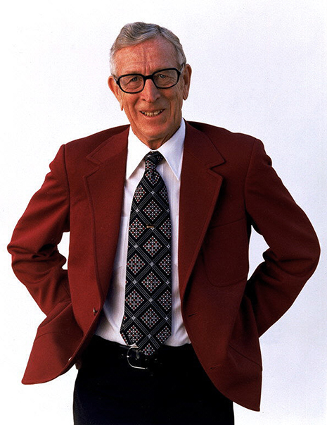 Wooden Color photo of John Wooden from the waist up, in a red suit jacket and grey patterned tie, from front.