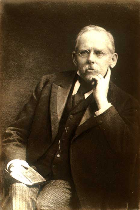 Black and white self-portrait photo of Jacob Riis, seated, wearing glasses and holding a book in his right hand.