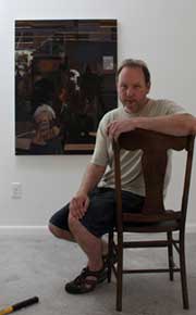 Color photo of artist Aaron Brown seated in a wooden chair in front of a painting.