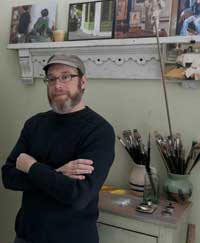 Color photo of artist Vincent Giarrano standing near a shelf of photographs and a table with paintbrushes