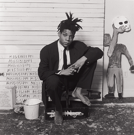 Black and white photo of Jean-Michel Basquiat in a black suit, crouching barefoot near artwork.