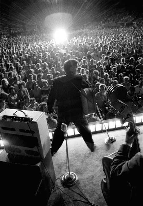 Black and white photo from of Elvis performing, taken from behind the stage, with spotlight on Elvis