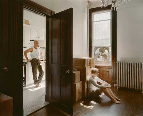 Photograph of man standing in kitchen, and woman in seperate room, sitting with packing boxes