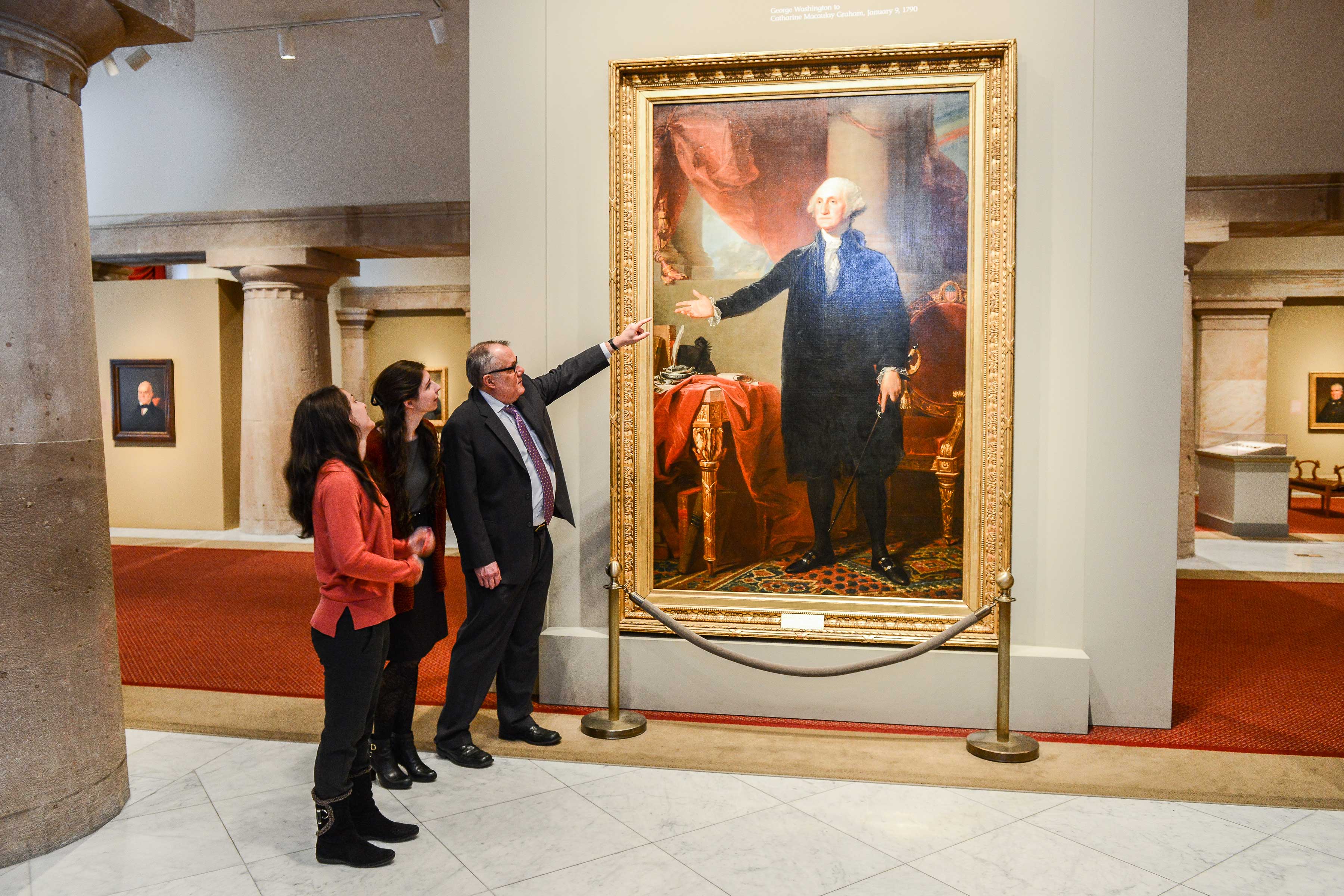 Picture of a man pointing towards a painted full-length portrait of George Washington 