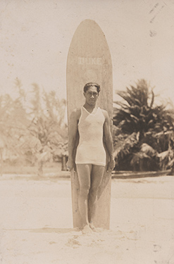 sepia toned full length photo of a man standing in front of a surfboard