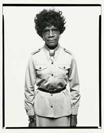 Black and white photo of a woman standing with glasses, short hair, a long jacket, and a belt