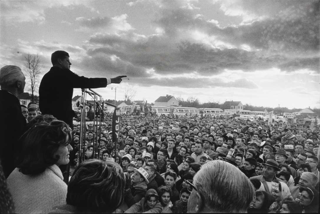 Black and white image of a man standing on a podium and pointing his finger at a very large crowd while speaking