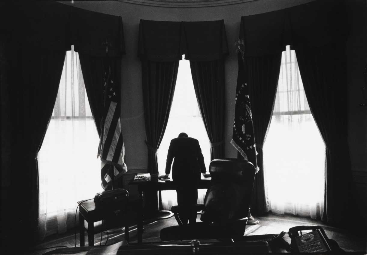 Black and white photo of the White House oval office with the sihouette of a man facing away from the camera in the frame of a window