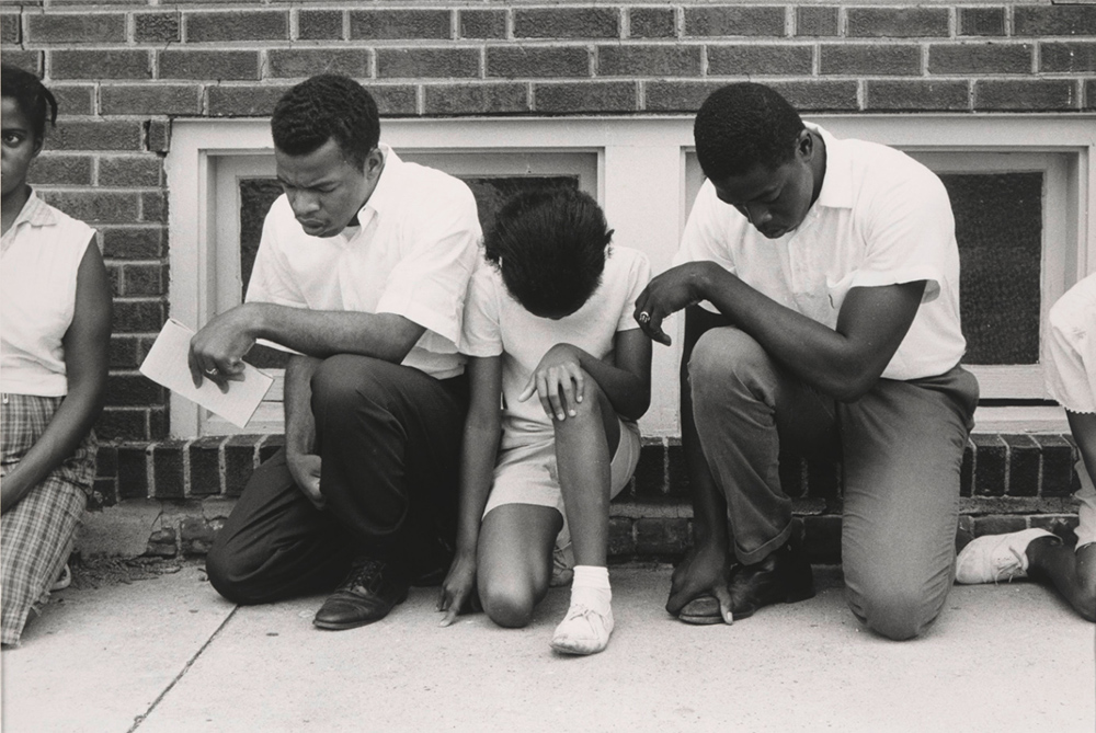 a group of African American activists kneeling on a sidewalk in prayer