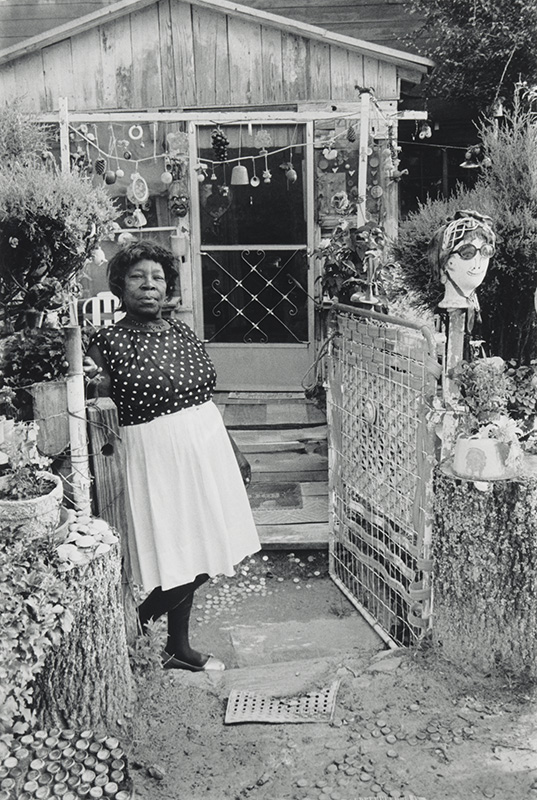 African American woman standing outside a small house structure surrounded by folk art
