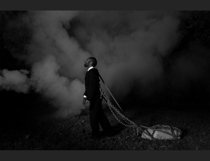 African American man in a suit dragging a large rock with ropes tied to him.