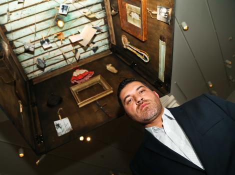 Man photographed from below. He is standing inside of a wooden box filled with photographs and other memorabilia.