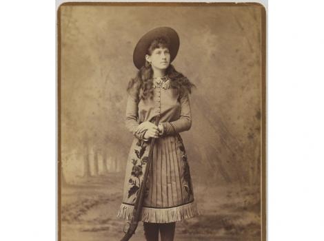 Photograph of a woman in western garb with a gun leaning against her leg