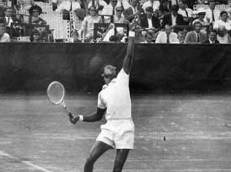Black and white photo of Arthur Ashe. He is tossing the ball for his serve, at a tennis match