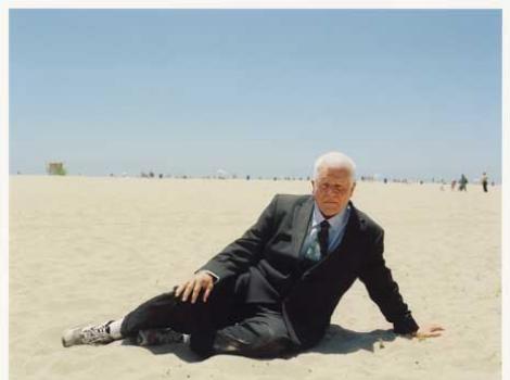 Color photo of an elderly man in a black business suit on a beach sitting in the sand.