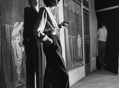Black and white photo of Lauren Bacall standing on a stage in front of a backdrop, with a cigarette in her left hand.
