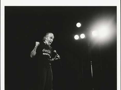 Black and white photo of George Carlin on stage wearing dark clothing and holding a microphone.