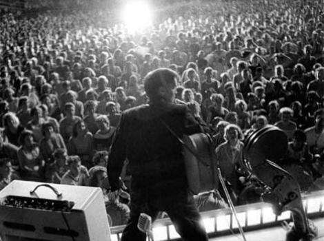 Black and white photo from of Elvis performing, taken from behind the stage, with spotlight on Elvis