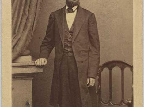 Black and white photo of Abraham Lincoln, standing next to a chair, with his right hand posed on a pedestal.