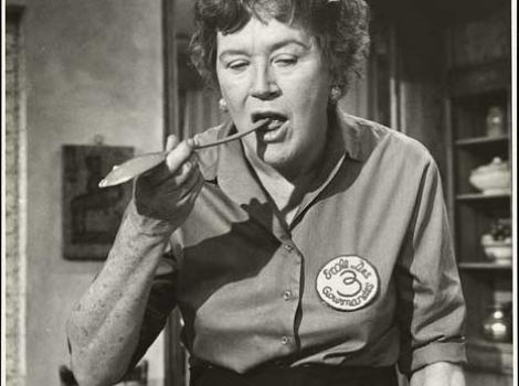 Black and white photo of Julia Child tasting in her kitchen with a wooden spoon