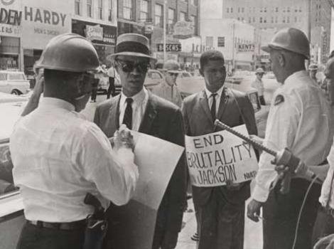 Black and white photo of Roy Wilkins and Medgar Evers, dressed in suits and being confronted by two police officers