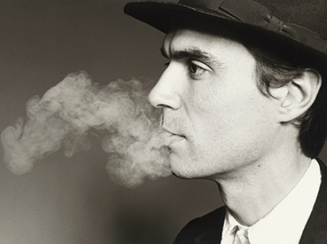 Black and white photo of David Byrne, smoking, wearing a dark suit and hat.
