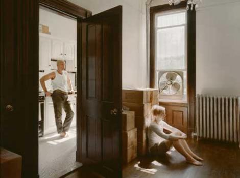 Photograph of man standing in kitchen, and woman in seperate room, sitting with packing boxes