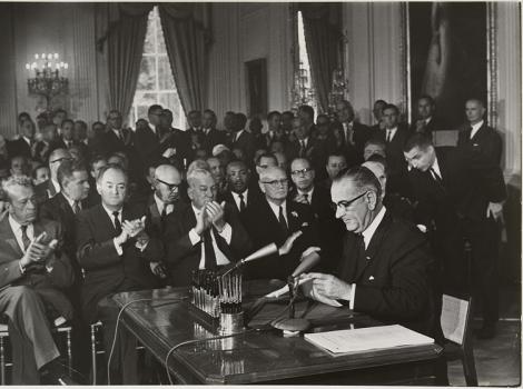 A black and white image of a man sitting at a desk signing a piece of paper with a lot of men clapping behind him