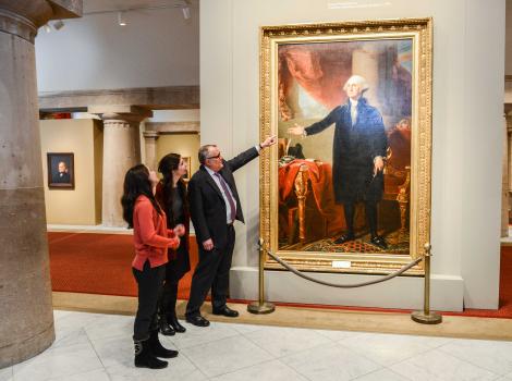 Picture of a man pointing towards a painted full-length portrait of George Washington 