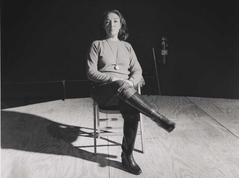 Black and white photo of a woman sitting cross-legged on a chair in the middle of a stage