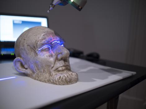 Small white plaster cast of a bearded man's face is scanned by a large laser that casts a blue light